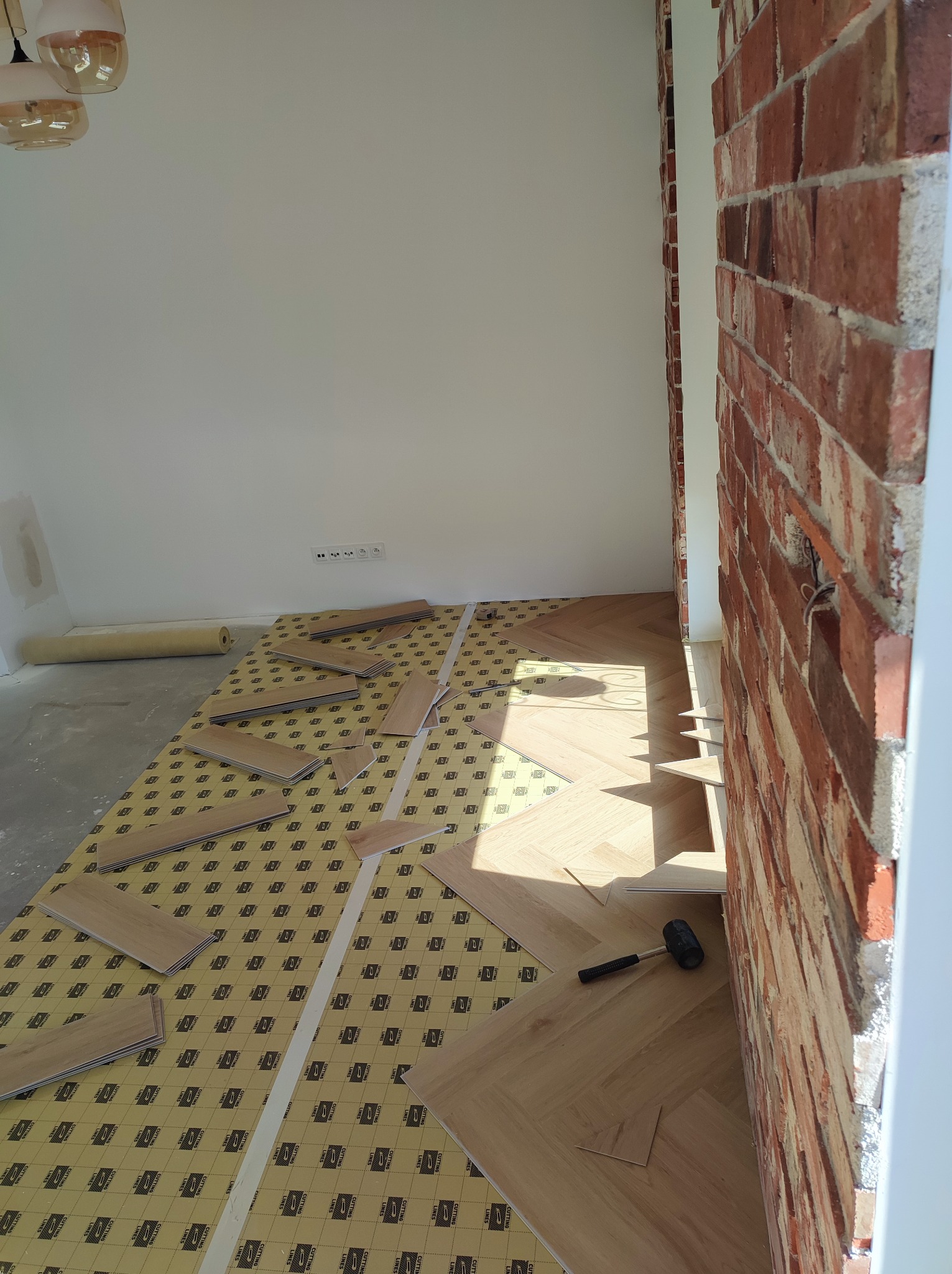 Układanie parkietu w jodełkę na podkładzie izolacyjnym, widoczny gumowy młotek i fragment ceglanej ściany w surowym stanie.