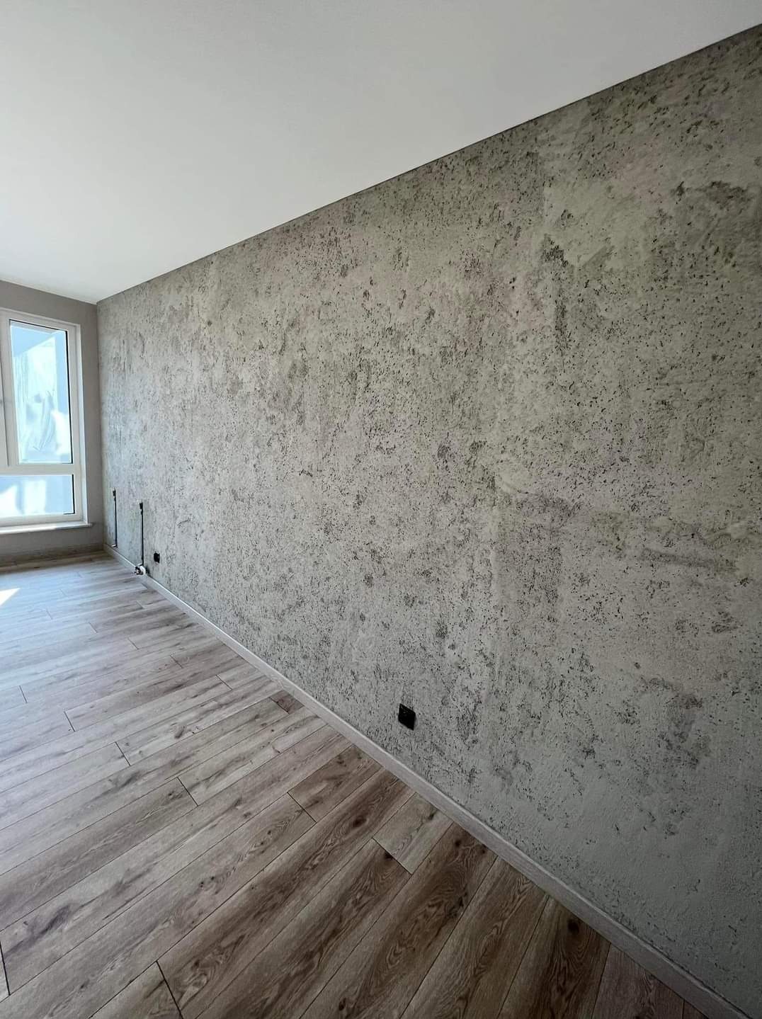 Ściana w pokoju wykończona dekoracyjną, szarą gładzią o nieregularnej fakturze, z widocznymi drewnopodobnymi panelami podłogowymi i oknem w tle.