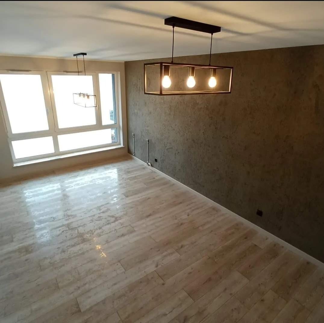Jasne panele podłogowe imitujące drewno w pustym pokoju z oknem i nowoczesnymi lampami wiszącymi, ściana pokryta dekoracyjnym tynkiem w odcieniach brązu.