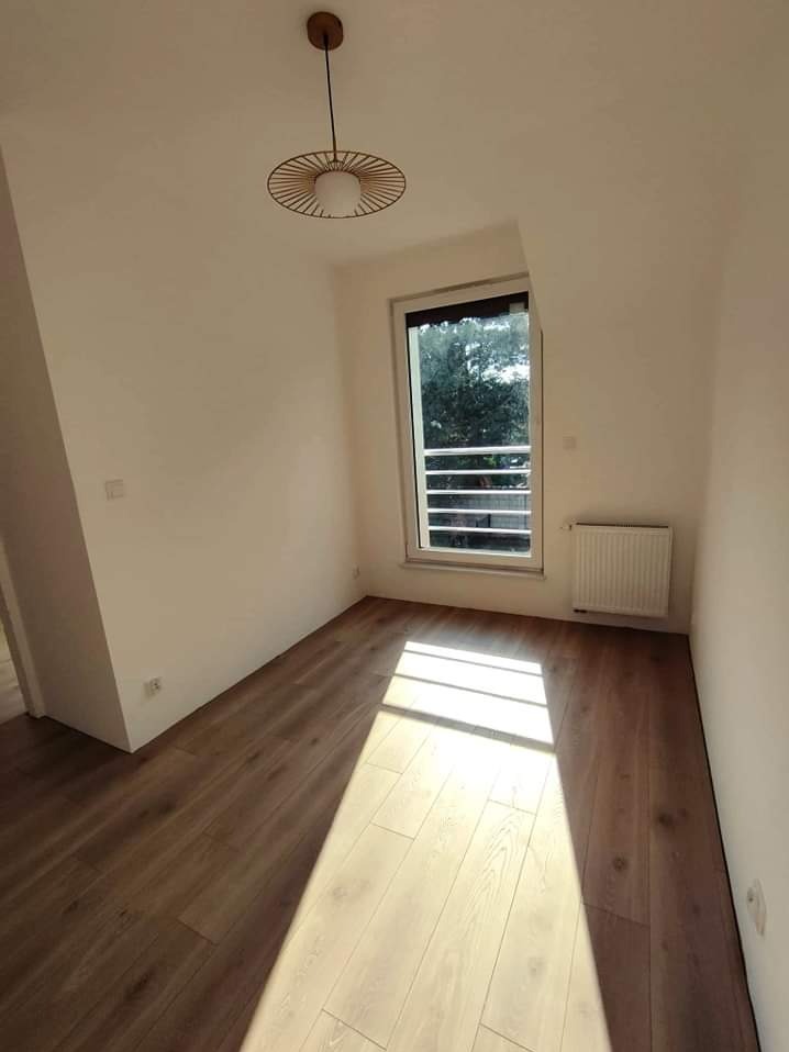 Jasne panele podłogowe w pustym pokoju z oknem i grzejnikiem, nowoczesna lampa sufitowa, ściany pomalowane na biało.