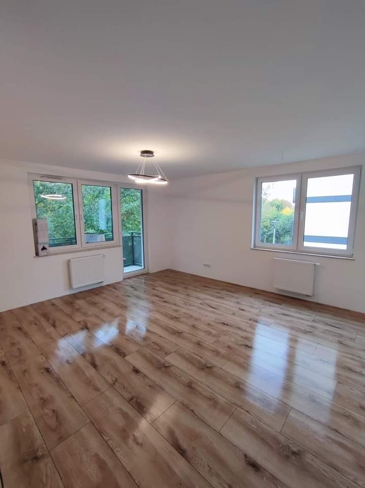 Jasne, puste pomieszczenie z nową podłogą z paneli imitujących drewno, białymi ścianami, grzejnikami pod oknami i nowoczesną lampą wiszącą.