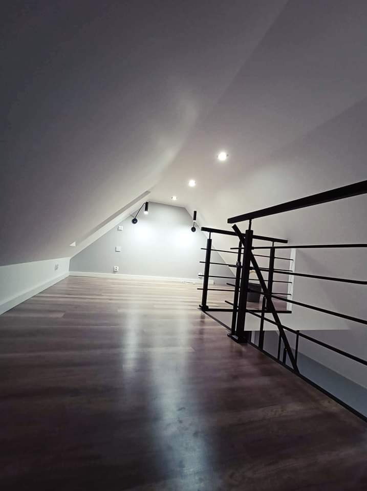 Poddasze po remoncie z nową, ciemną podłogą panelową i pomalowanymi na szaro ścianami, oświetlone punktowymi lampami sufitowymi i z czarną metalową balustradą.