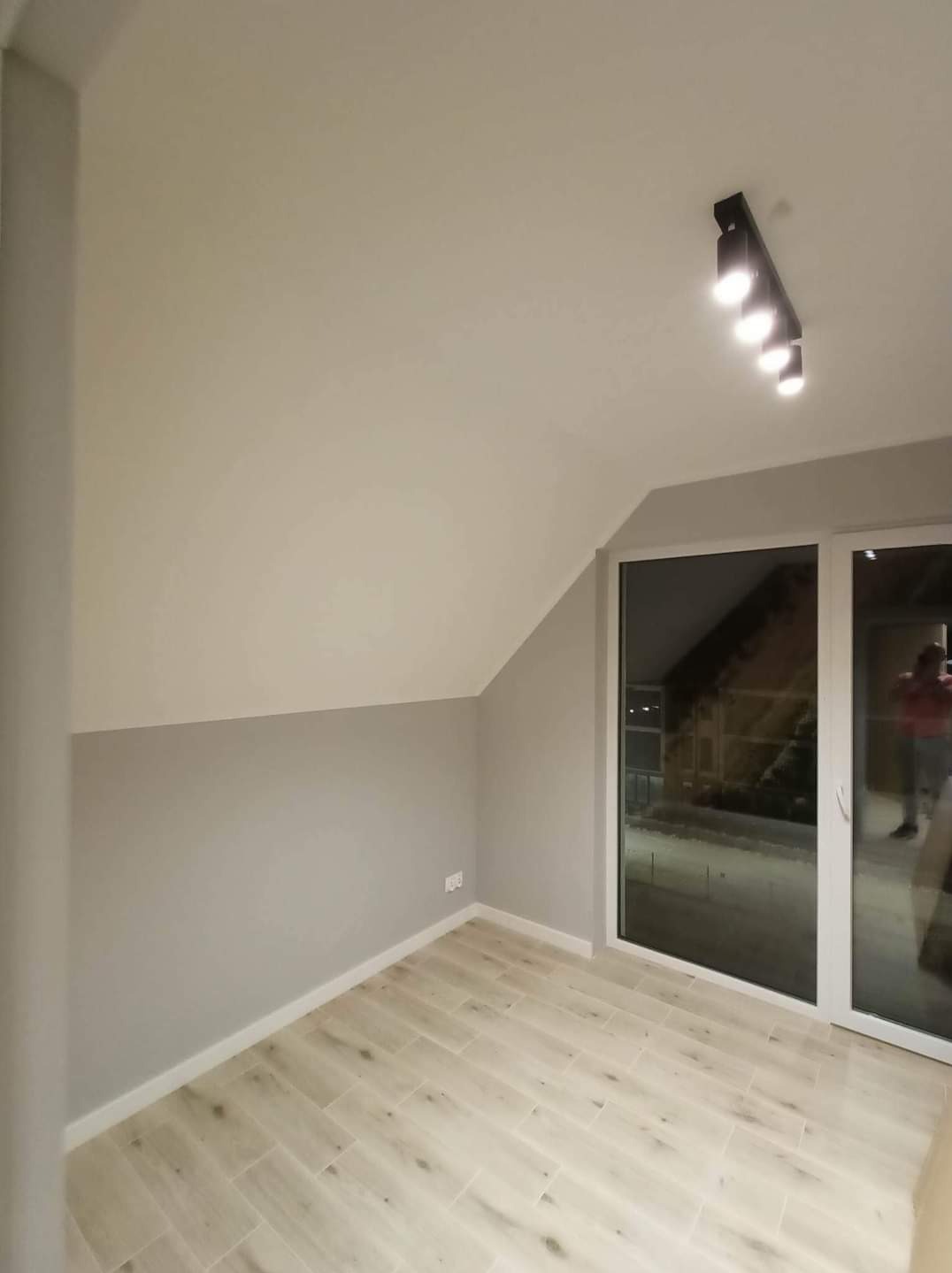 Pokój na poddaszu po remoncie: ściany pomalowane na dwa odcienie szarości, jasna podłoga z paneli, czarne oświetlenie sufitowe i duże okno balkonowe z widokiem na zewnątrz.