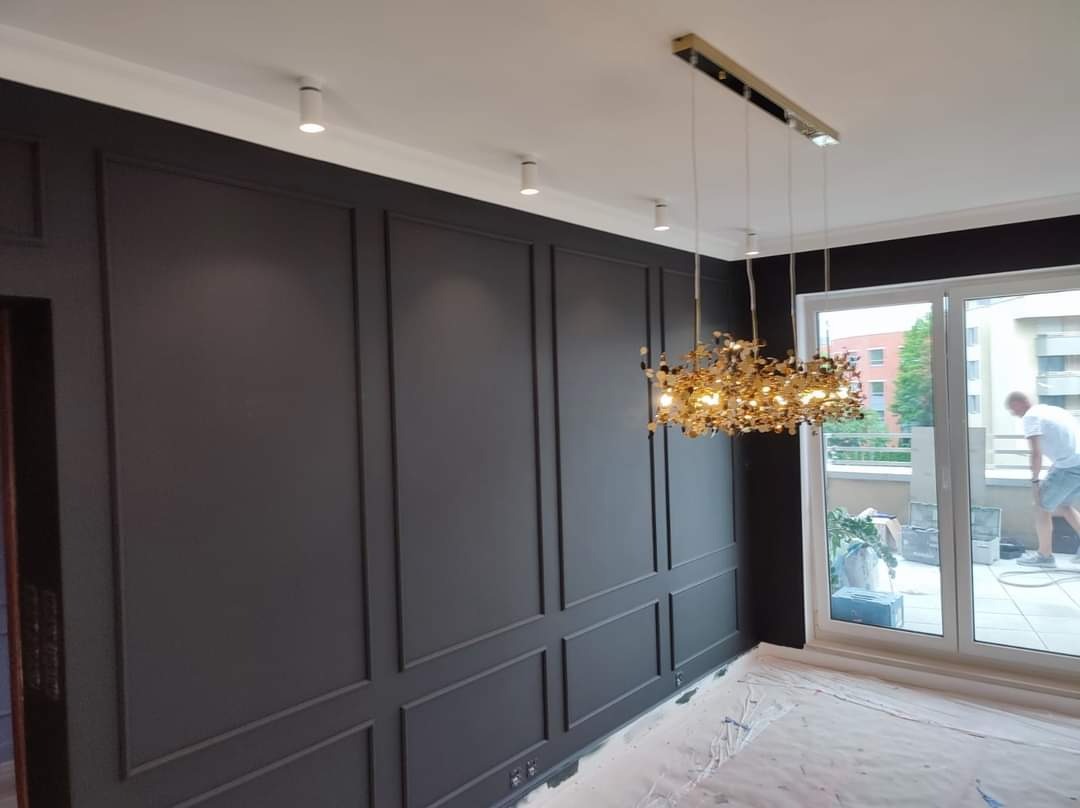 Eleganckie wnętrze po malowaniu, ciemnoszara ściana z dekoracyjnymi panelami, nowoczesny złoty żyrandol, podłoga zabezpieczona folią malarską, widok na balkon z pracującym mężczyzną.
