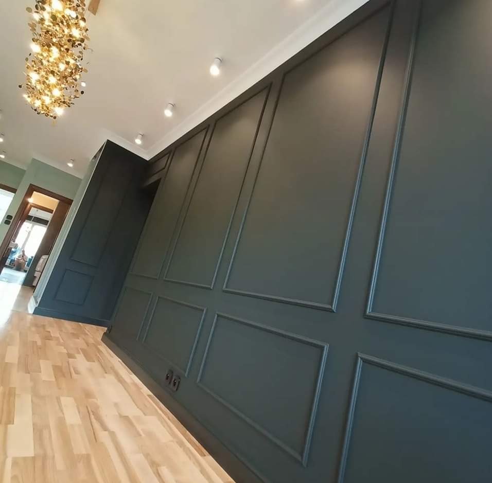 Elegancko wykończone wnętrze z ciemnozielonymi panelami ściennymi z dekoracyjnymi listwami, jasną drewnianą podłogą i designerskim żyrandolem.