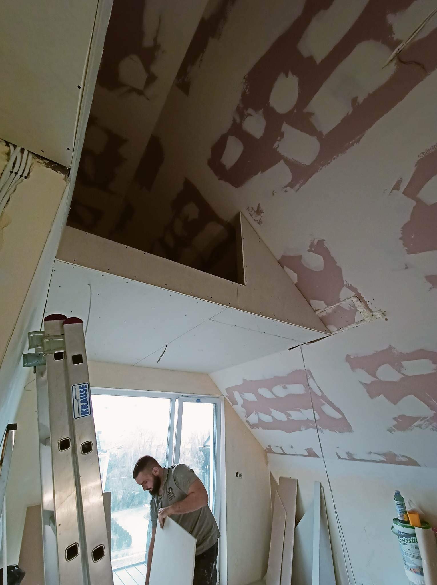 Prace wykończeniowe poddasza: montaż płyt gipsowo-kartonowych na skosach, widoczny pracownik z płytą, drabina Krause i fragment okna.
