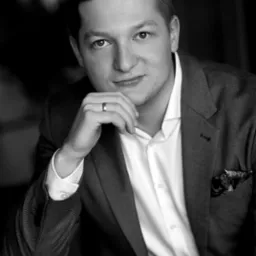 Portret czarno-biały mężczyzny w marynarce i białej koszuli, z ręką podpierającą brodę, na palcu widoczna obrączka, w tle rozmyte wnętrze.