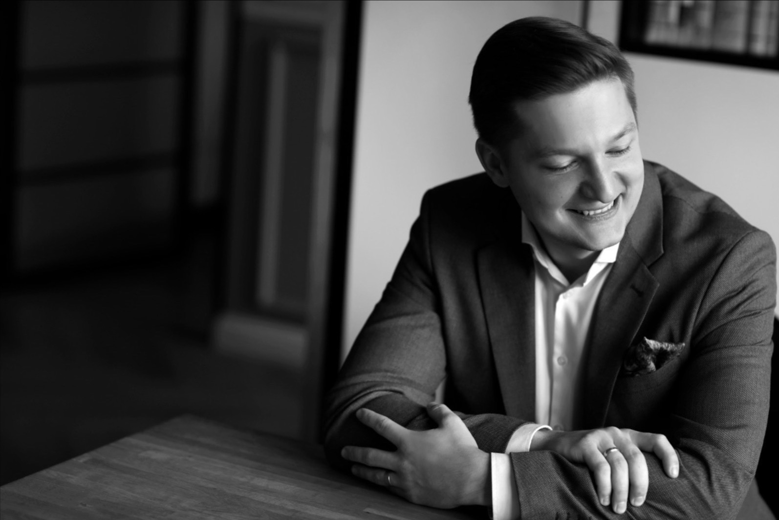 Portret uśmiechniętego mężczyzny w garniturze, siedzącego przy drewnianym stole, w czarno-białej tonacji.