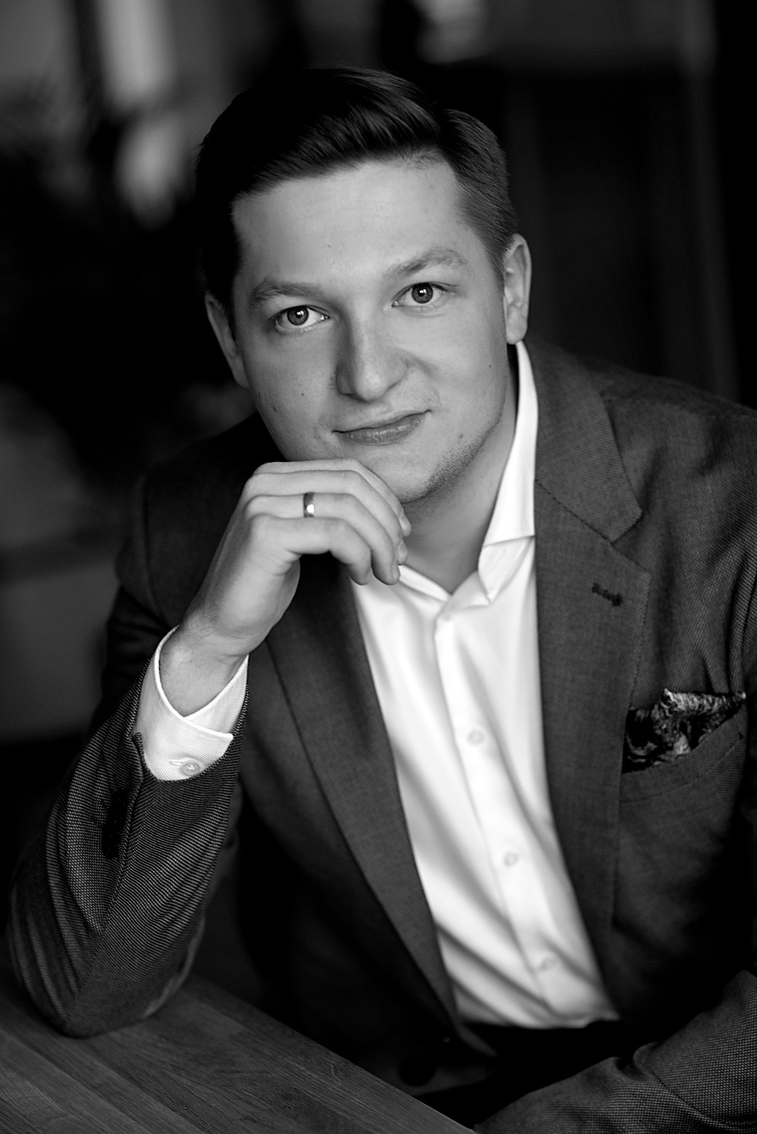 Portret czarno-biały mężczyzny w marynarce i białej koszuli, z ręką podpierającą brodę, na palcu widoczna obrączka, w tle rozmyte wnętrze.