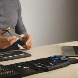 Serwisant delikatnie odłącza ekran uszkodzonego smartfona, narzędzia rozłożone na stole, w tle otwarty laptop.