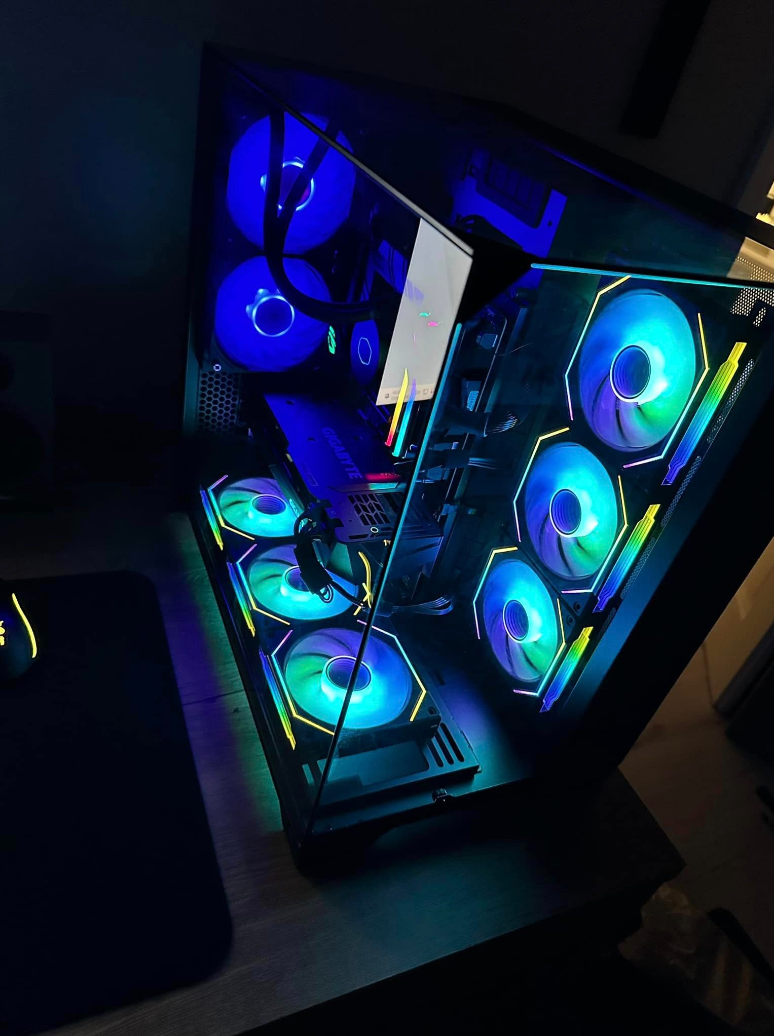 Przezroczysta obudowa komputera stacjonarnego z widocznymi wentylatorami RGB, kartą graficzną Gigabyte i chłodzeniem wodnym, ustawiona na biurku obok myszki komputerowej.