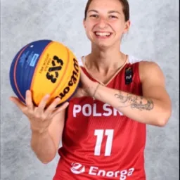 Uśmiechnięta kobieta w czerwonej koszulce sportowej z napisem POLSKA trzyma piłkę do koszykówki 3x3 Wilson, widoczny tatuaż na ramieniu.