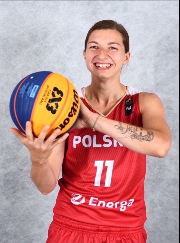 Uśmiechnięta kobieta w czerwonej koszulce sportowej z napisem POLSKA trzyma piłkę do koszykówki 3x3 Wilson, widoczny tatuaż na ramieniu.