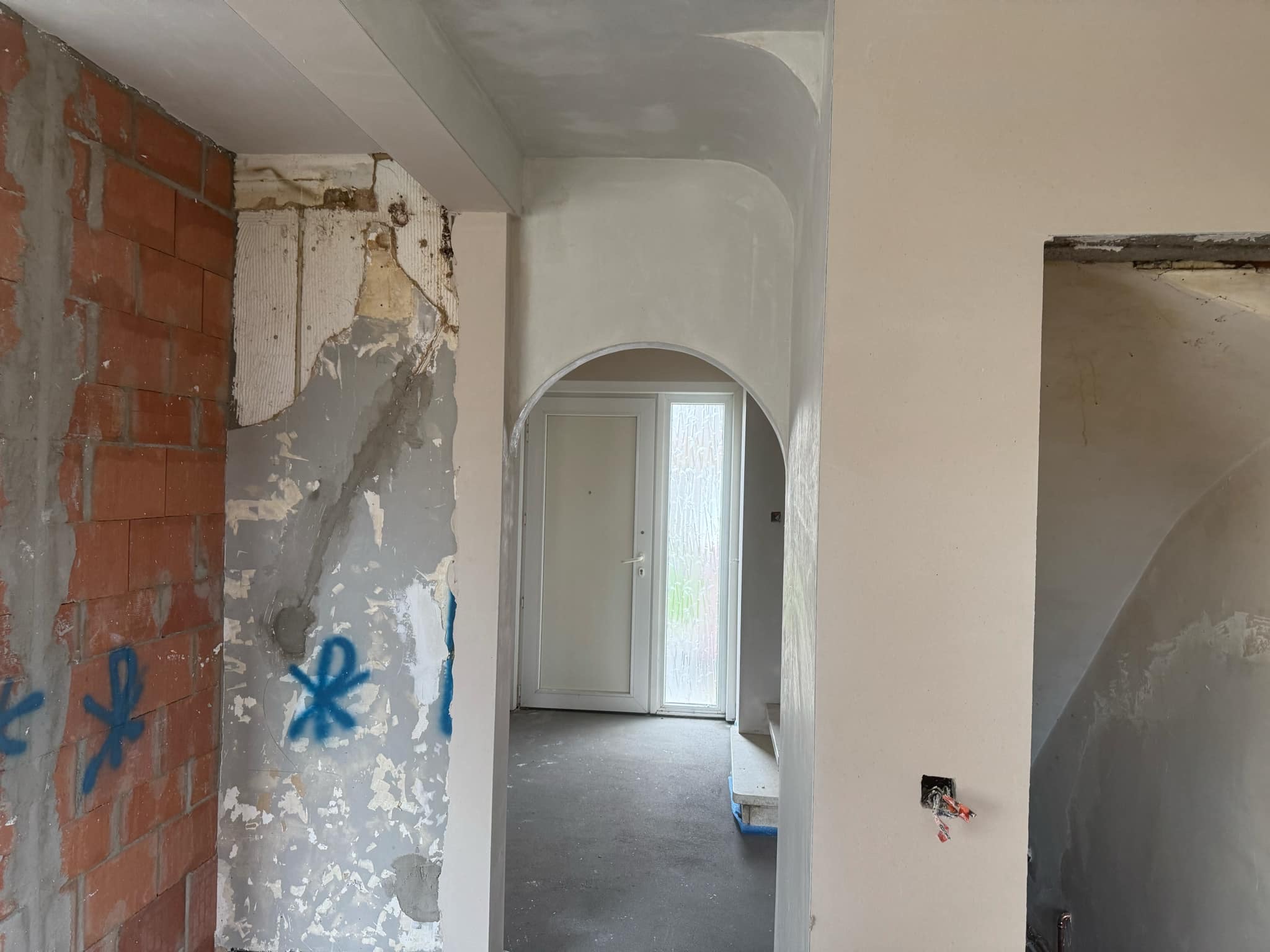 Wnętrze w trakcie remontu: surowe ściany z cegły i resztkami tapet kontrastują z gładkimi, szpachlowanymi łukami przejść. Widoczne instalacje elektryczne i drzwi w tle.