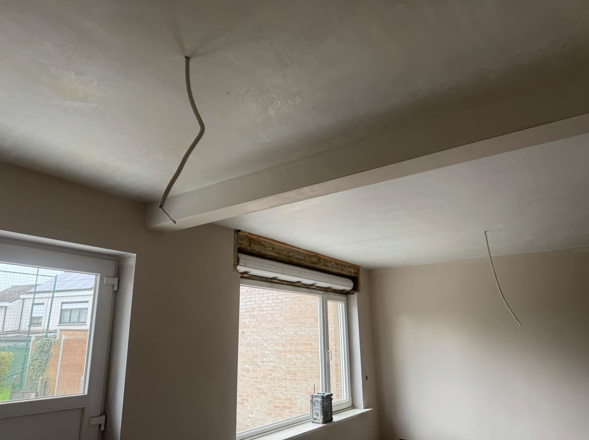 Wnętrze po szpachlowaniu: gładka, jasna ściana i sufit z wystającym przewodem, okno z roletą. Widok na ceglany budynek zza okna. Świeżo wykończone powierzchnie.