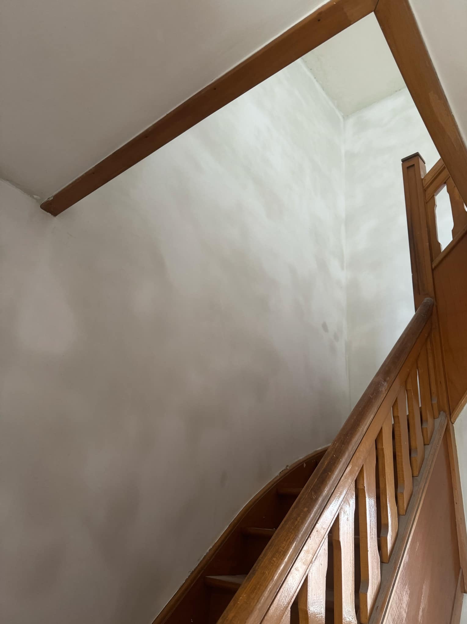 Ściana po szpachlowaniu w klatce schodowej z drewnianą balustradą. Widoczne nierówności i ślady po szpachli, przygotowane do malowania. Ujęcie z dołu.
