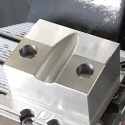 Obrabiany aluminiowy blok z dwoma otworami i wyfrezowanym rowkiem, zamocowany na stole frezarki CNC, widoczne ślady chłodziwa.