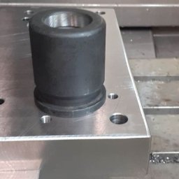 Eryk Gibasiwicz - Czarny, cylindryczny element z otworem, zamocowany na aluminiowej płycie z otworami, leżącej na stole frezarki CNC w Sulmierzycach, z widocznymi wiórami metalowymi.