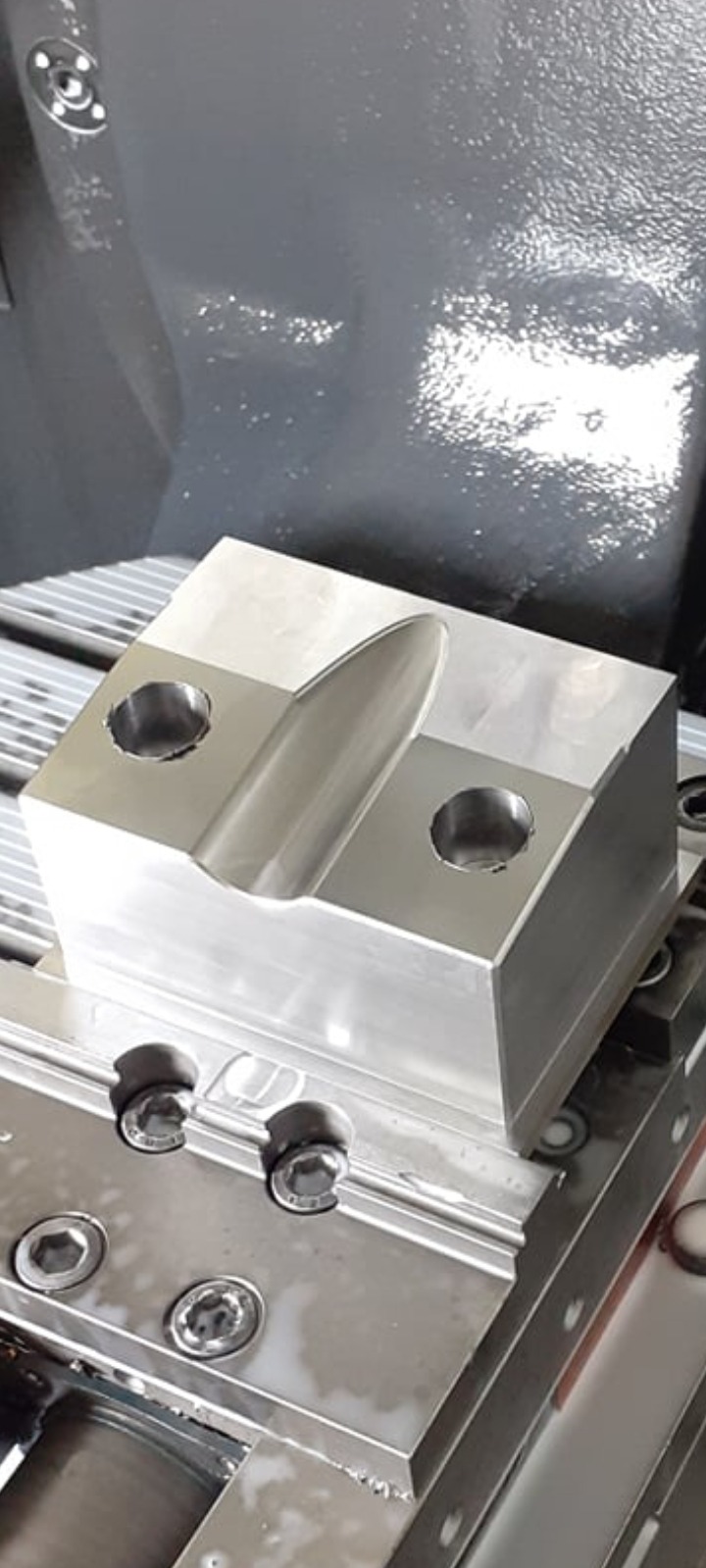 Obrabiany aluminiowy blok z dwoma otworami i wyfrezowanym rowkiem, zamocowany na stole frezarki CNC, widoczne ślady chłodziwa.