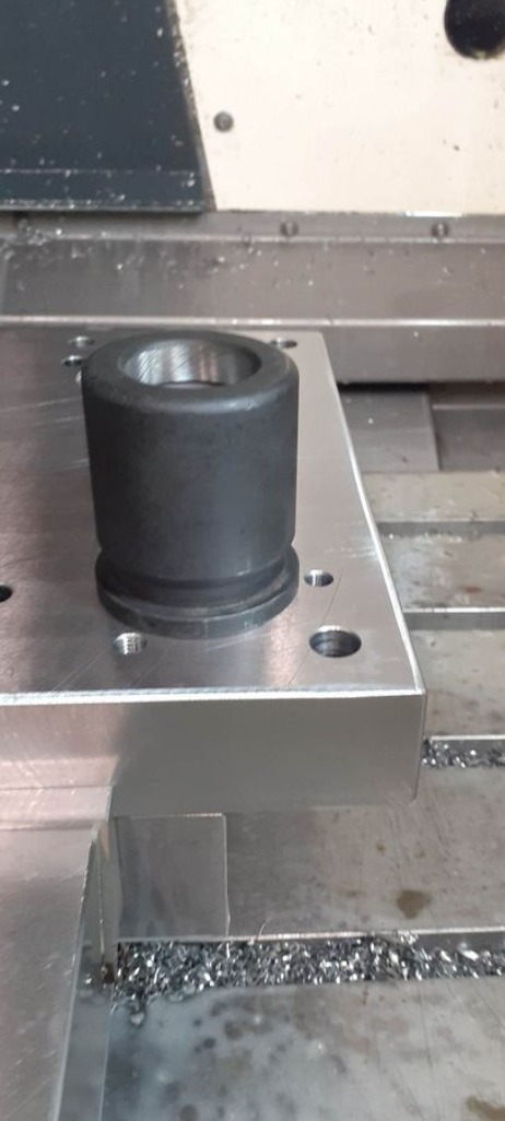 Czarny, cylindryczny element z otworem, zamocowany na aluminiowej płycie z otworami, leżącej na stole frezarki CNC w Sulmierzycach, z widocznymi wiórami metalowymi.