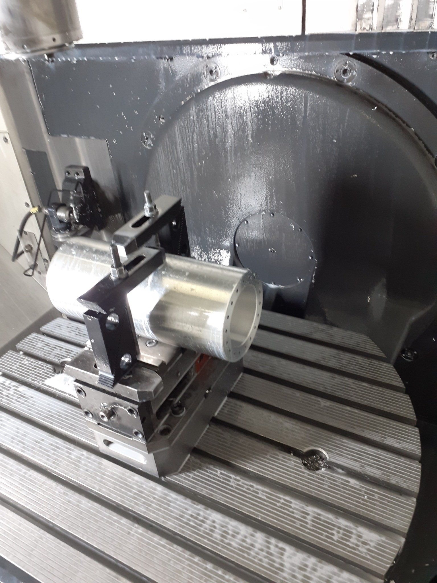 Cylindryczny element aluminiowy mocowany w uchwycie na stole obrotowym frezarki CNC, widoczne ślady chłodziwa.