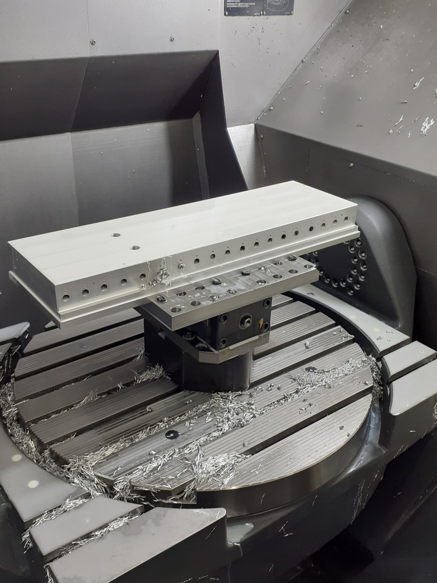 Obrabiany aluminiowy element z rzędem otworów na stole frezarki CNC, wióry po obróbce.