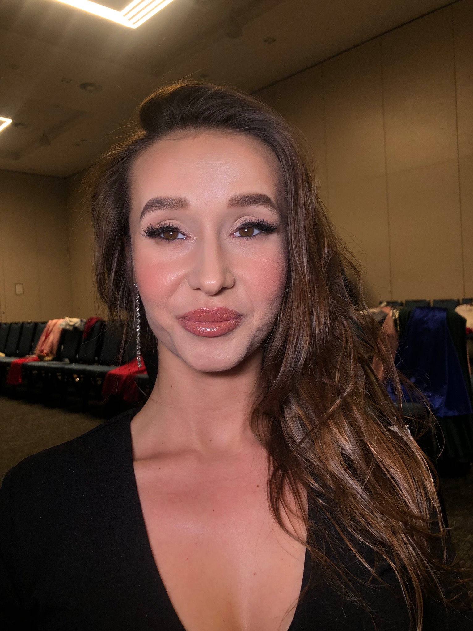 Portret kobiety z delikatnym, dziennym upiększeniem, długimi kolczykami i brązowymi włosami na tle garderoby teatralnej.