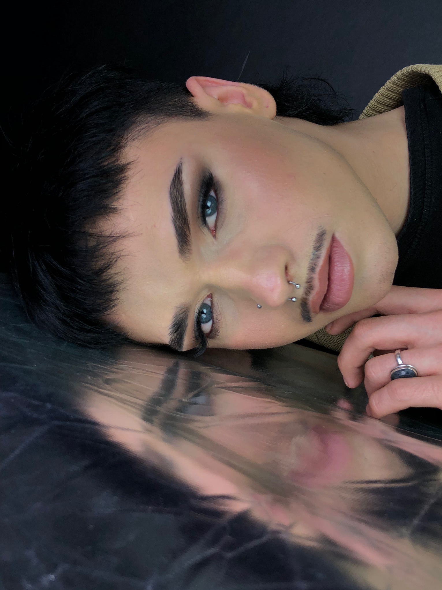 Portret osoby z delikatnym wąsikiem, piercingiem w nosie i wyrazistym makijażem oczu, leżącej na srebrzystej powierzchni, z pierścieniem na palcu.