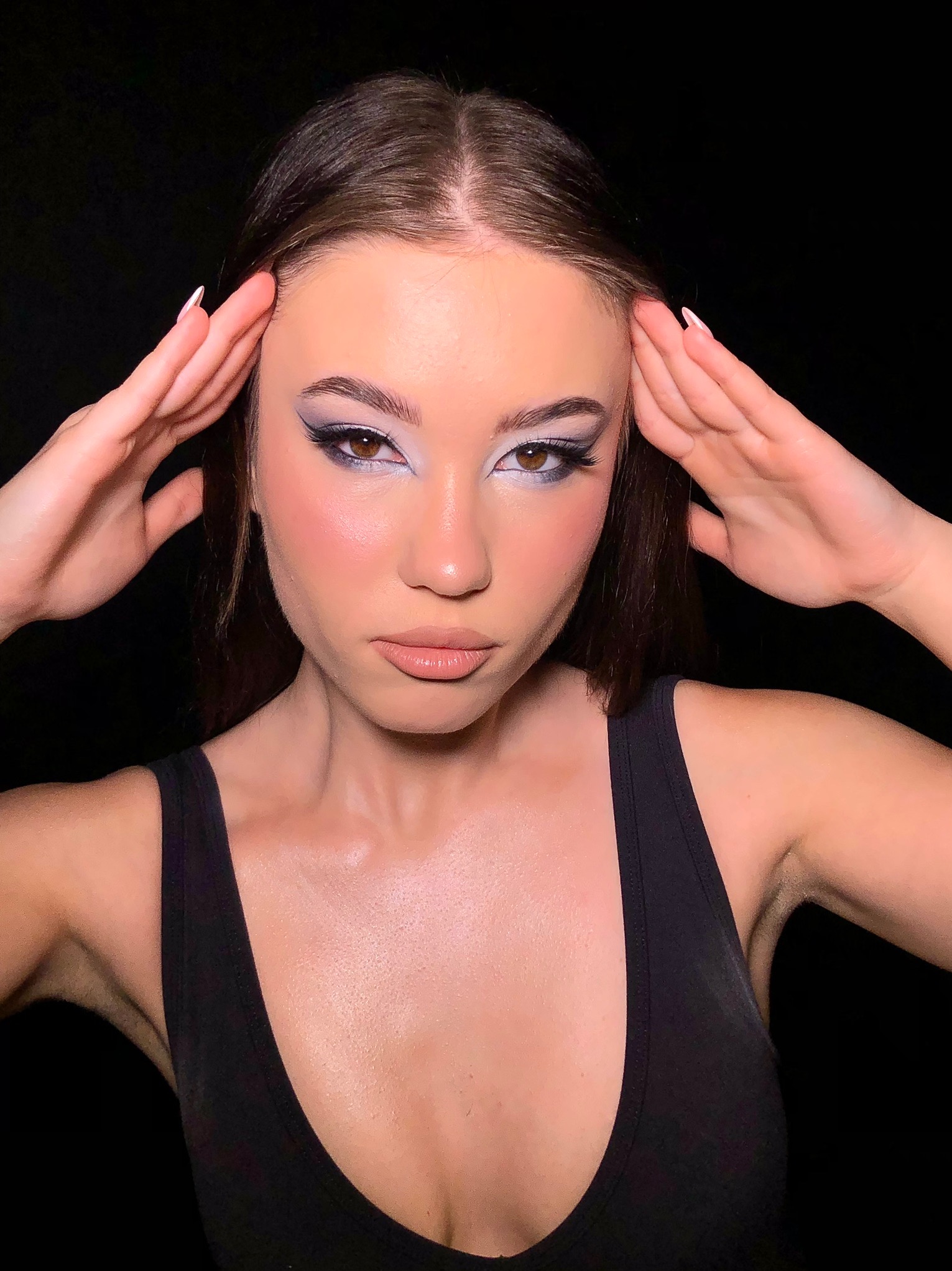 Portret kobiety z profesjonalnym, srebrzysto-szarym cieniowaniem powiek i wyraźną kreską, dłonie uniesione do góry, ciemne tło.