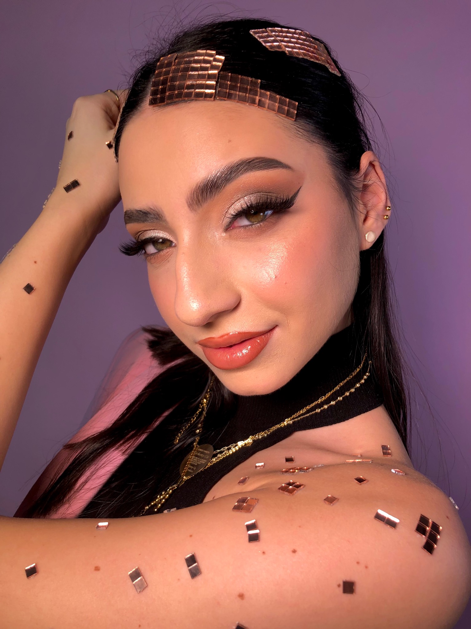 Portret kobiety z błyszczącym, srebrzystym cieniem na powiekach, podkreślonymi brwiami, sztucznymi rzęsami i błyszczykiem na ustach, ozdobiona kwadratowymi, miedzianymi aplikacjami na włosach...