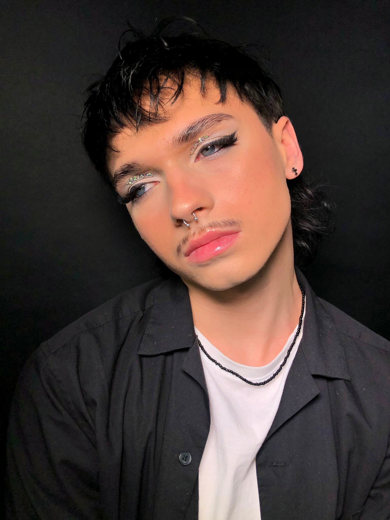 Osoba z ozdobnym, błyszczącym cieniem do powiek, mocno wytuszowanymi rzęsami, błyszczącymi ustami i kolczykiem w nosie na czarnym tle.