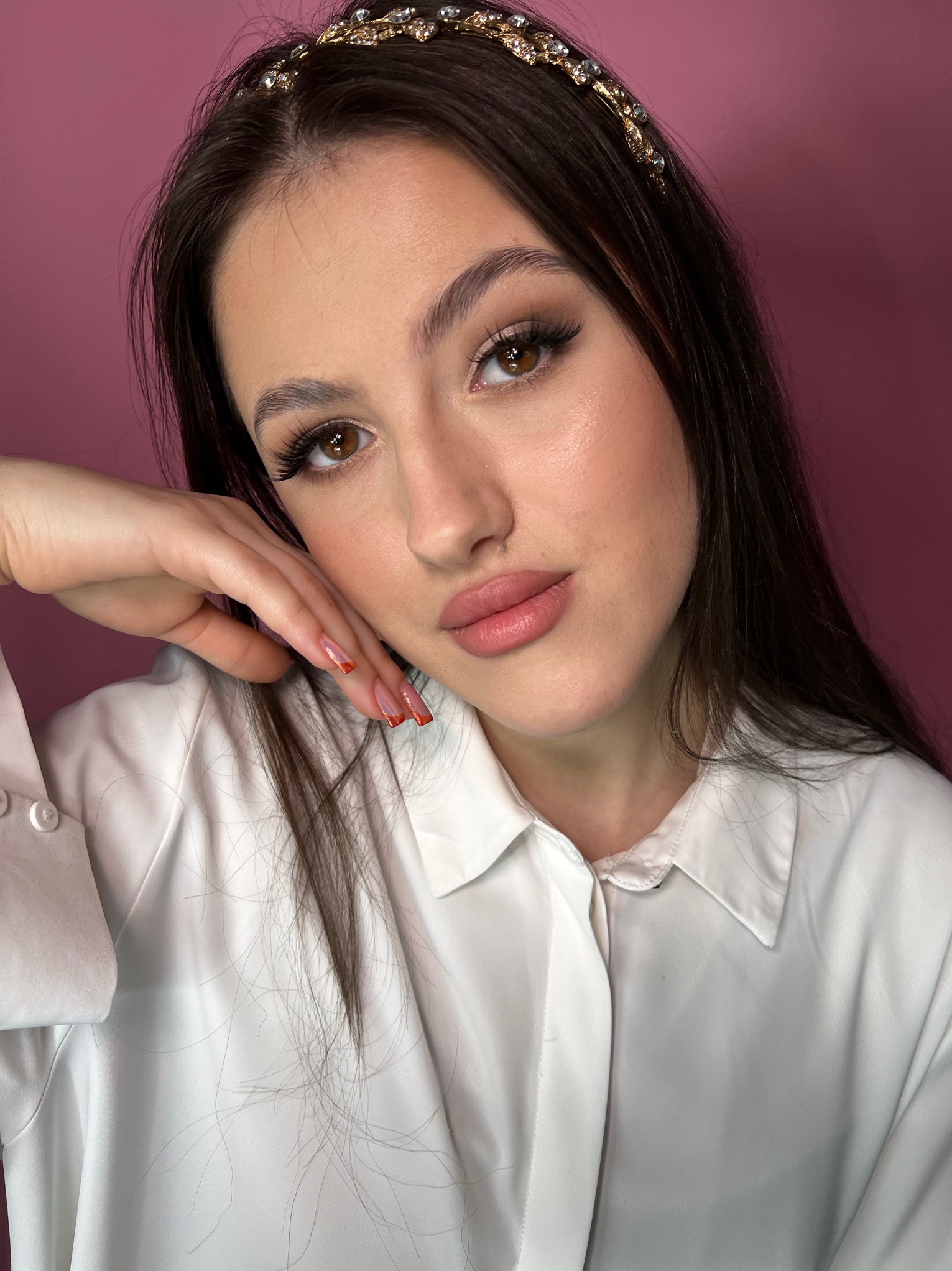 Portret brunetki z delikatnym, dziennym make-upem, ozdobną opaską na włosach i manicure hybrydowym z pomarańczowymi końcówkami na tle różowego studia.