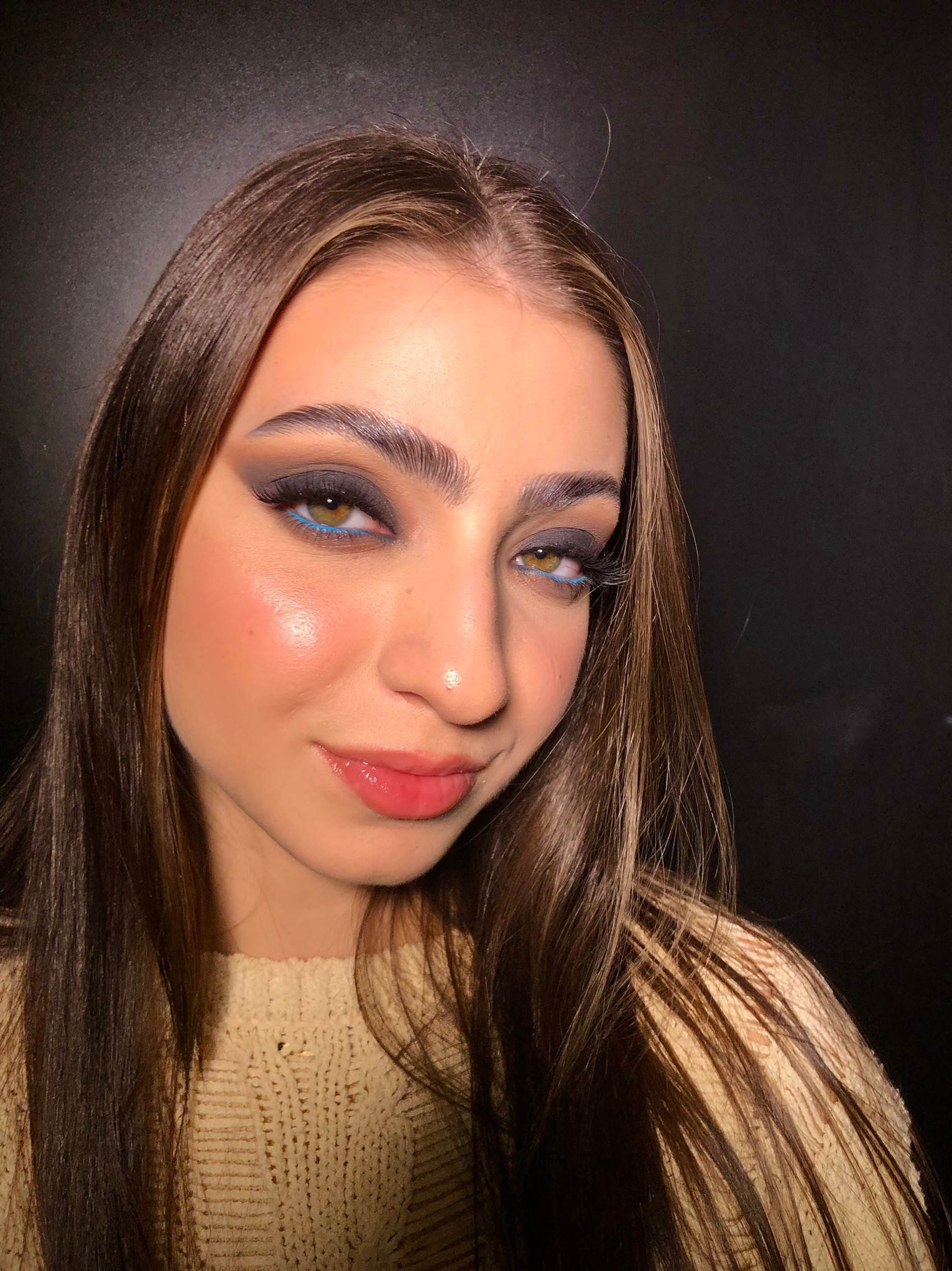 Portret kobiety z intensywnym, wieczorowym w odcieniach szarości i niebieskiego, błyszczącego pigmentu na powiekach, podkreślonymi brwiami i błyszczącymi ustami na tle czarnego tła.