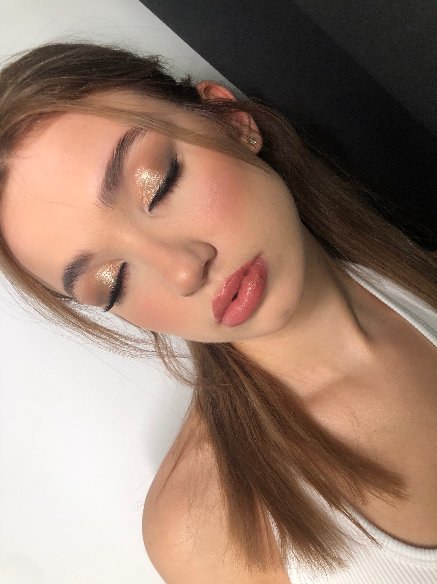 Portret młodej kobiety z zamkniętymi oczami, prezentujący złoty, błyszczący cień do powiek i delikatny róż na policzkach, z widocznym kolczykiem w uchu i naturalnym odcieniem ust.