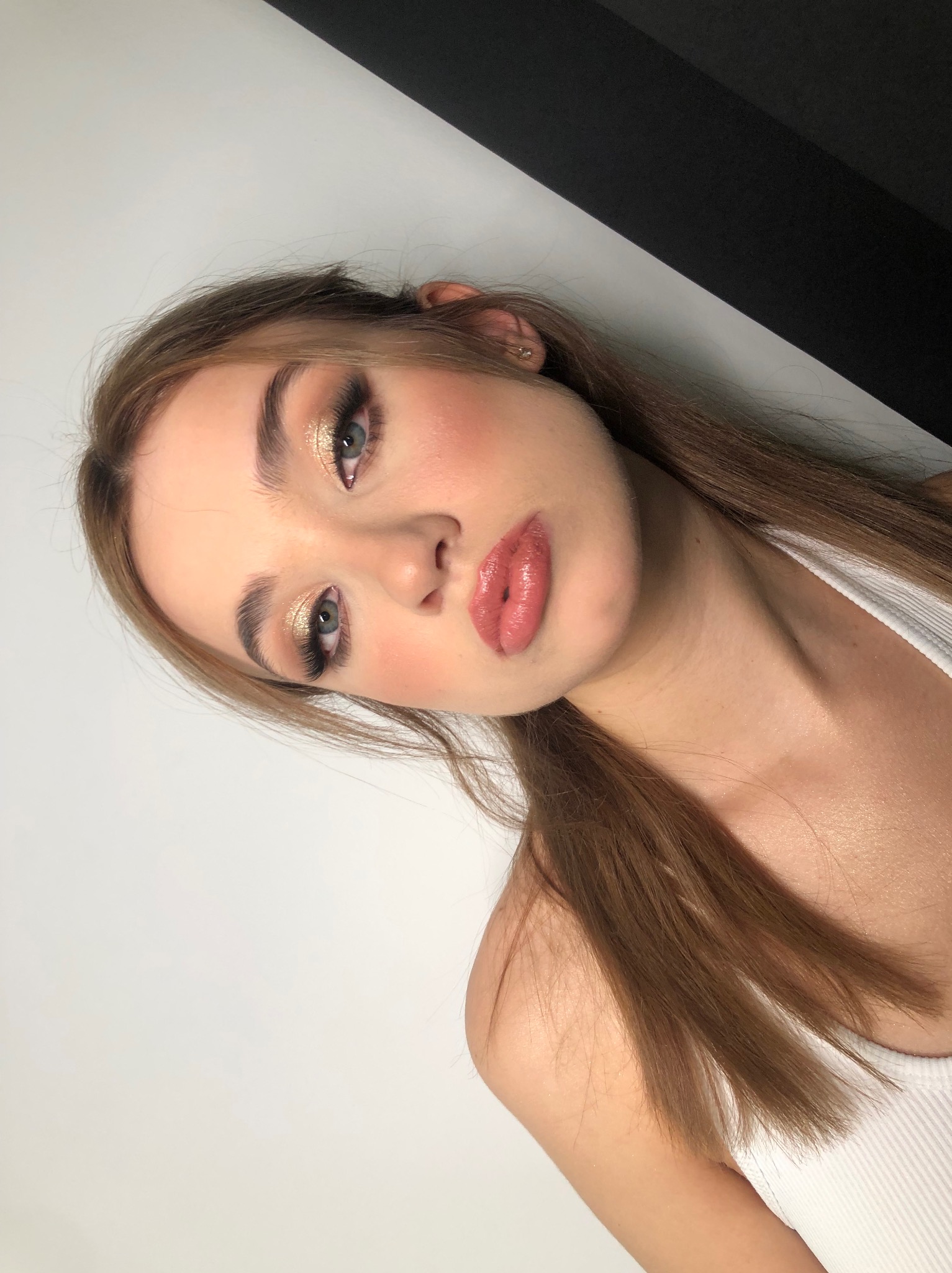 Portret młodej kobiety z profesjonalnym makijażem: błyszczący cień na powiekach, podkreślone rzęsy, róż na policzkach i błyszczące usta w odcieniu nude.