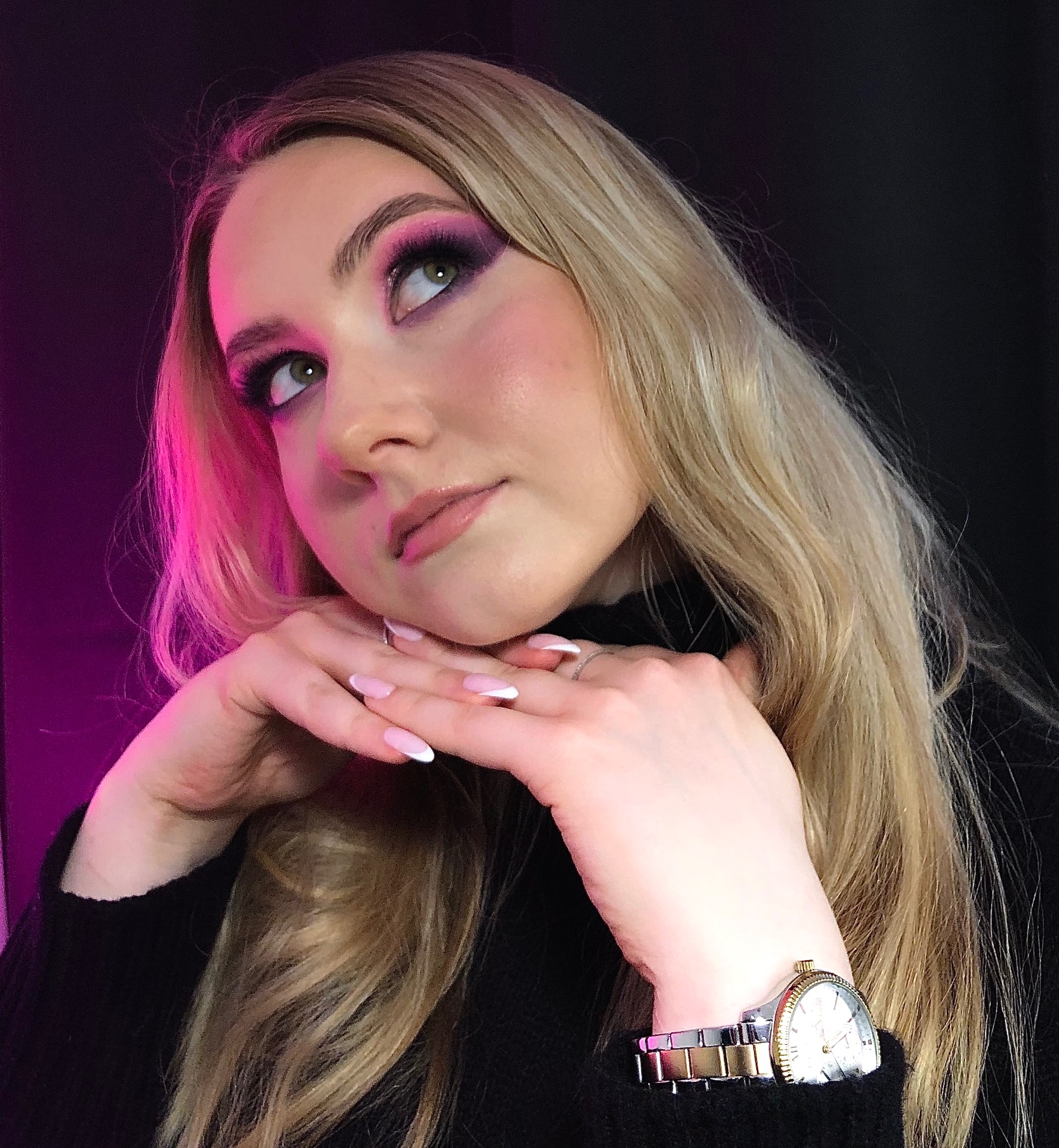 Portret kobiety z intensywnym, różowo-fioletowym cieniowaniem powiek, jasnymi włosami i delikatnym manicure w stylu french, pozującej z dłońmi pod brodą, w czarnym swetrze i z zegarkiem...