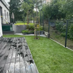 Nowoczesny ogród w Gdyni po deszczu: ciemne deski tarasu z kroplami wody, świeżo założony trawnik, młode drzewka z podporami, rabaty kwiatowe i szara siatka ogrodzeniowa.