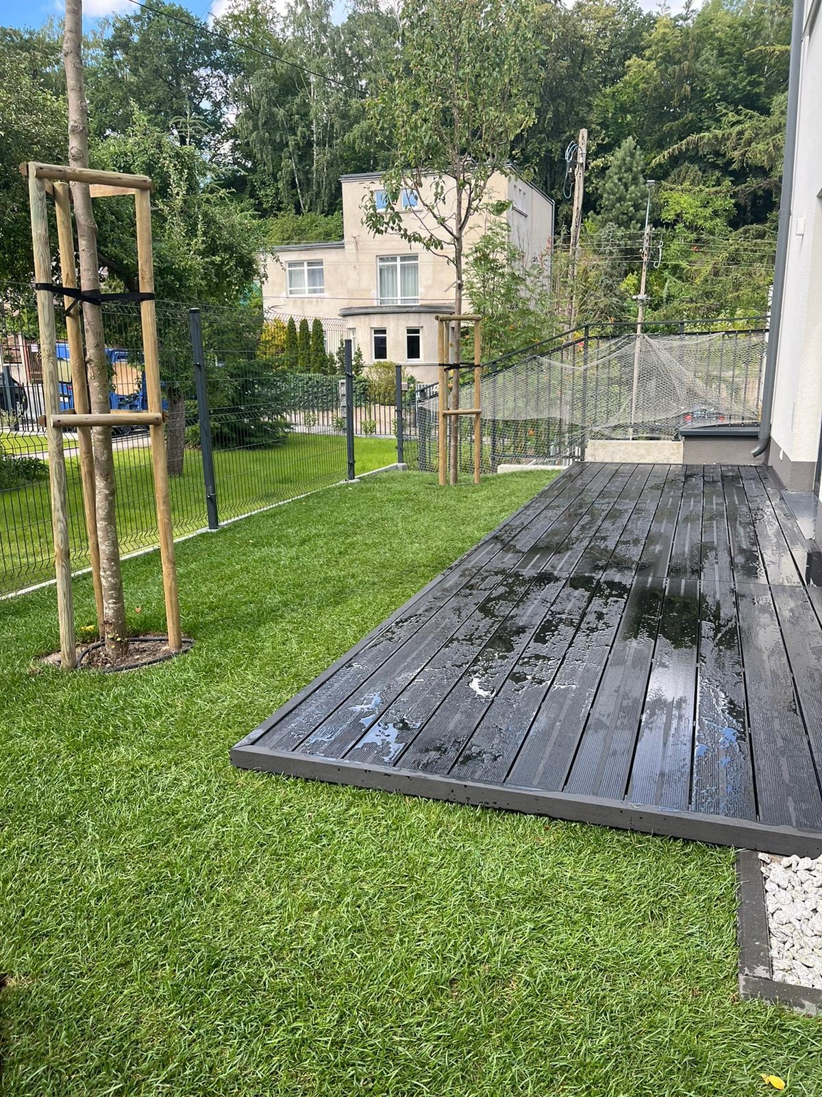 Świeżo założony trawnik z rolki, widoczne drzewka zabezpieczone drewnianą konstrukcją, mokry, drewniany taras przylegający do budynku, siatka ogrodzeniowa w tle.