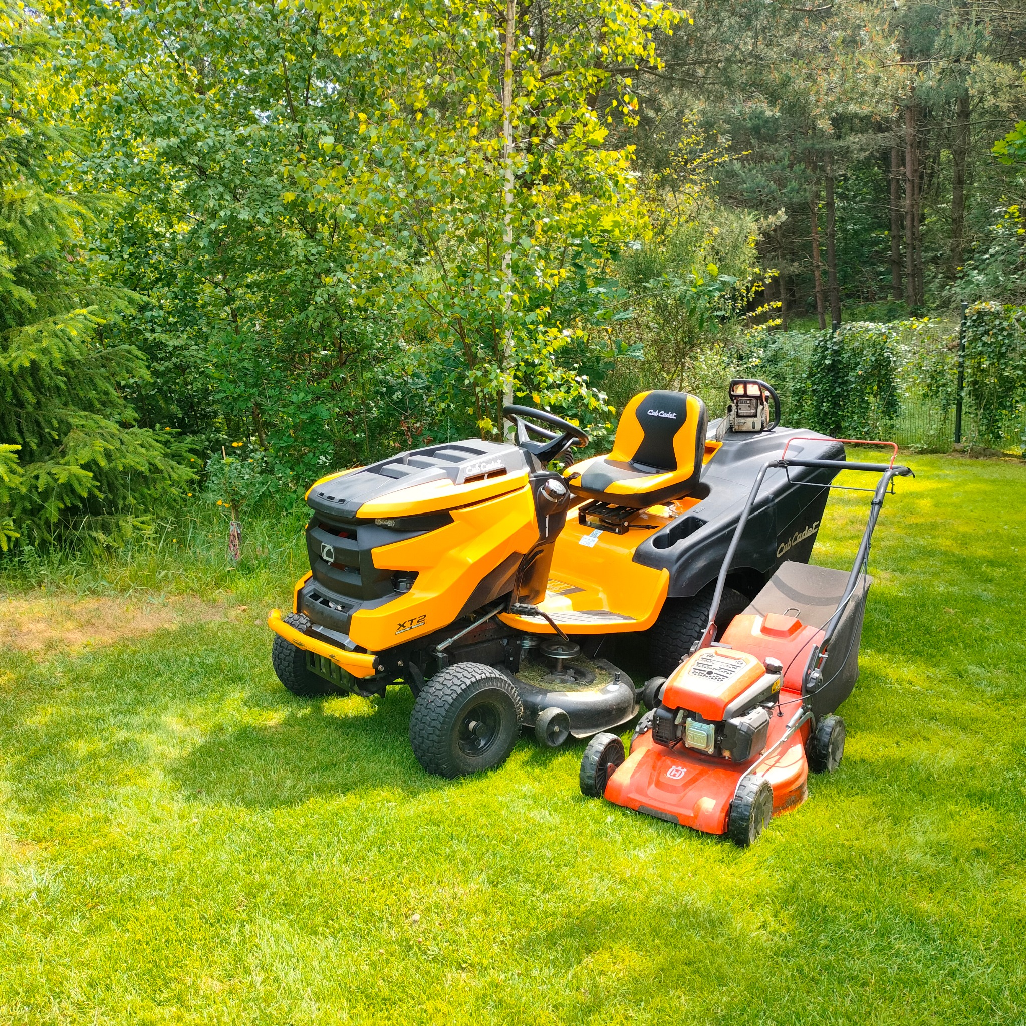 Żółty traktorek do koszenia trawy Cub Cadet XT2 i pomarańczowa kosiarka Husqvarna stoją na świeżo skoszonej trawie przed drzewami.