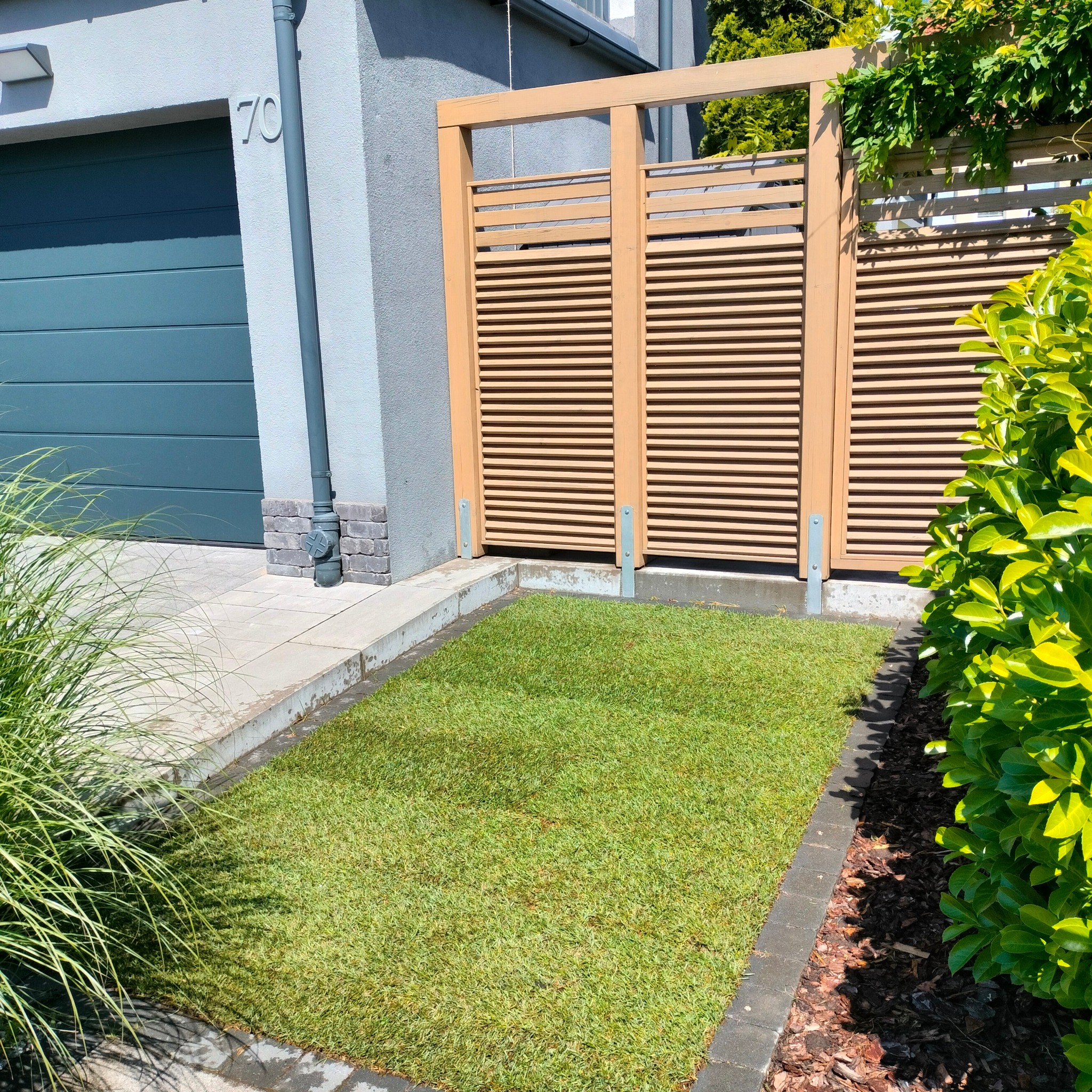 Świeżo ułożona trawa z rolki przed domem z szarą elewacją i drewnianym parawanem, widoczne obrzeża z kostki brukowej i fragment zieleni ozdobnej.