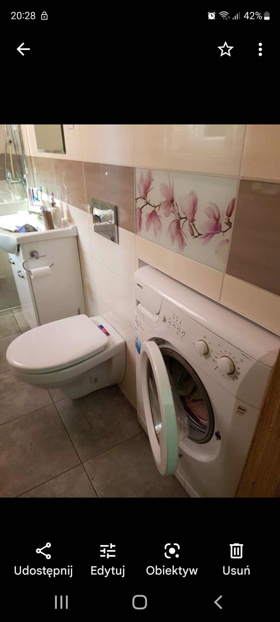 Ukończona łazienka z białą ceramiką, wiszącą toaletą i pralką Beko z otwartymi drzwiami, płytki z motywem magnolii na ścianie.