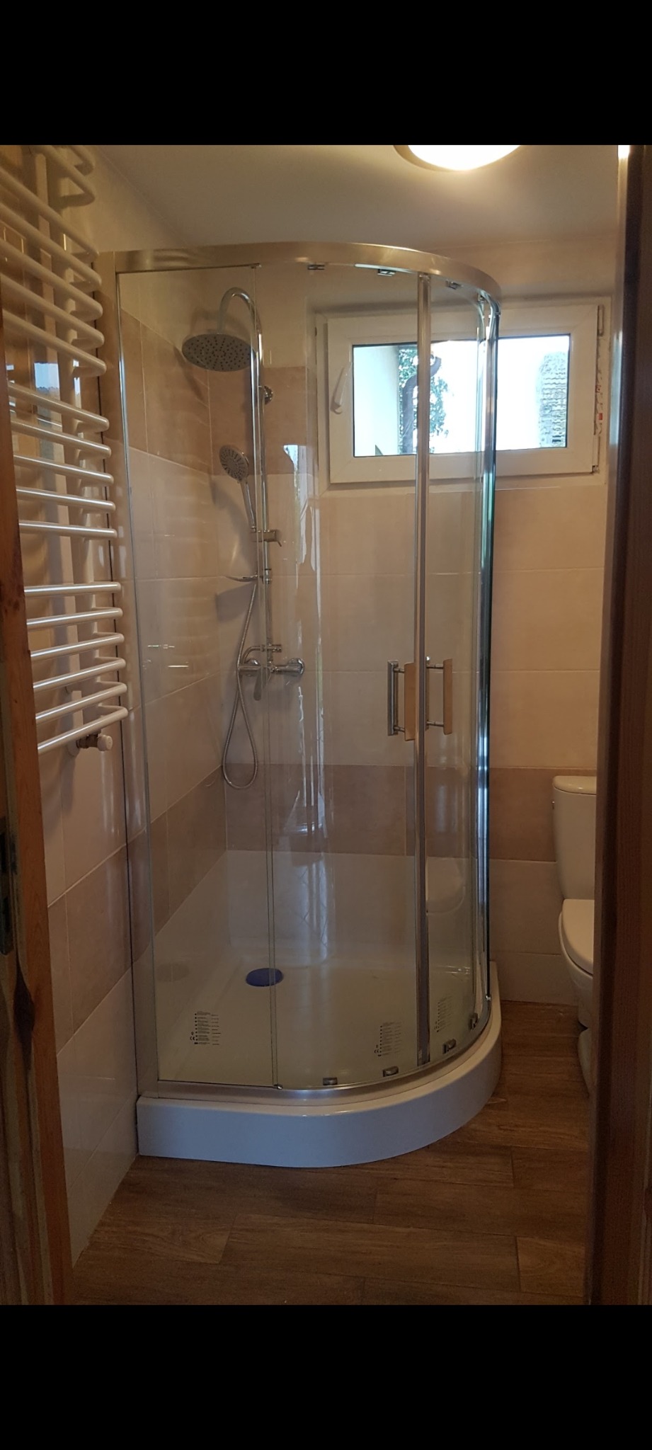 Nowoczesna kabina prysznicowa z hydromasażem, chromowanymi elementami i szklanymi drzwiami w łazience z beżowymi płytkami i drewnopodobną podłogą, widok zza otwartych drzwi.