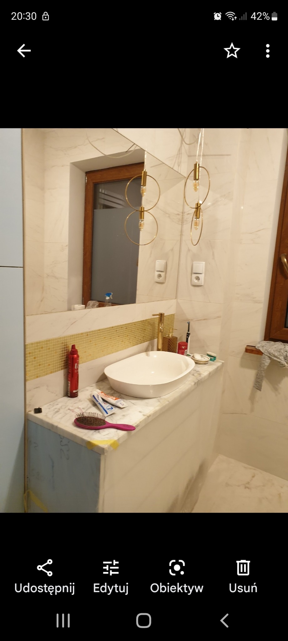 Nowoczesna łazienka z marmurową umywalką nablatową, złotymi akcentami w armaturze i oświetleniu, częściowo wykończona z widocznymi niedoróbkami na obudowie umywalki.