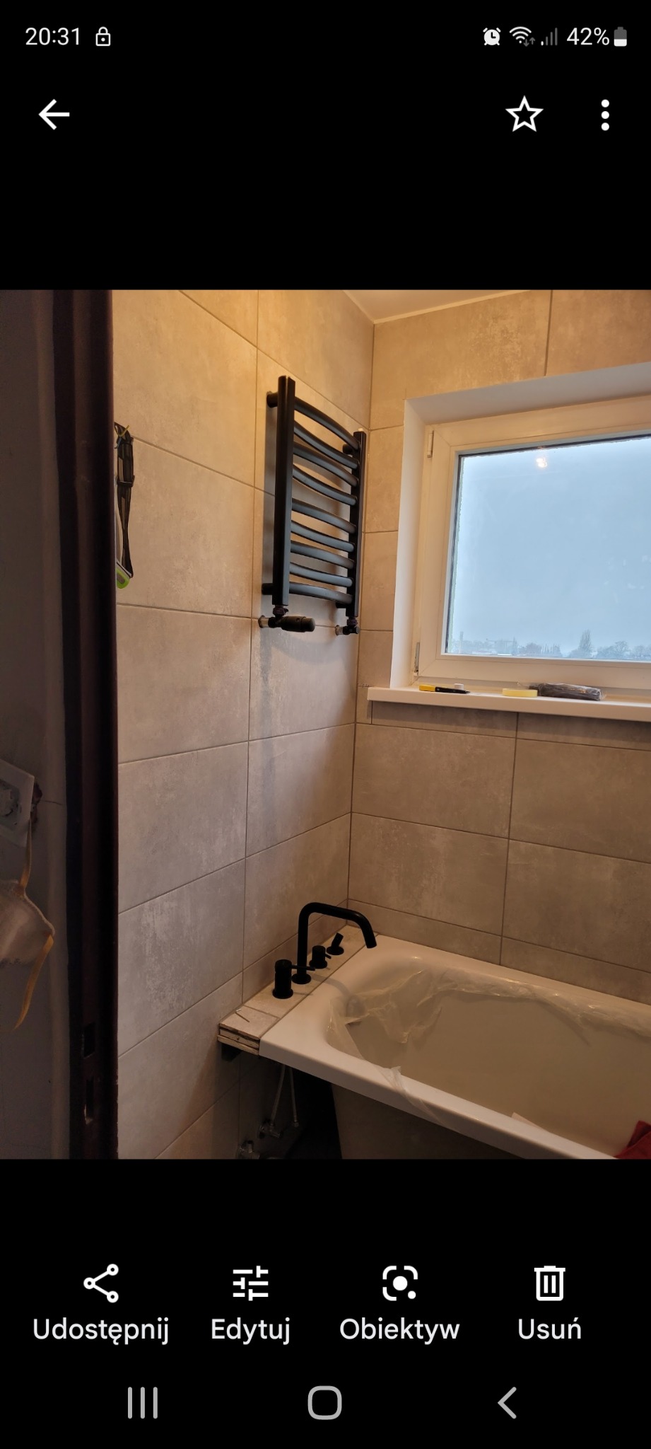 Nowa, czarna armatura łazienkowa: bateria wannowa i grzejnik drabinkowy na tle beżowych płytek, wanna zabezpieczona folią ochronną, widok na okno z pochmurnym niebem.