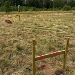 Prace geodezyjne w terenie: tyczka geodezyjna, niwelator na statywie i drewniane paliki z oznaczeniami wysokości na zarośniętej działce, w tle drzewa.