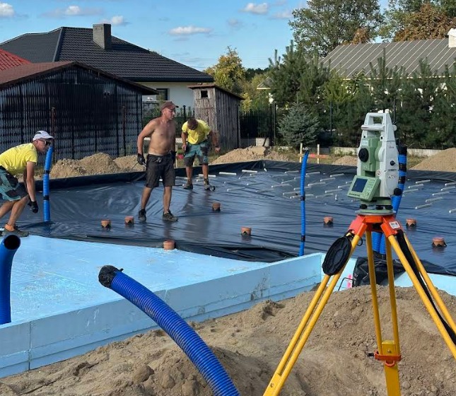 Prace przygotowawcze pod budowę: niwelacja terenu z użyciem niwelatora optycznego. Widoczni pracownicy i fundamenty w tle.