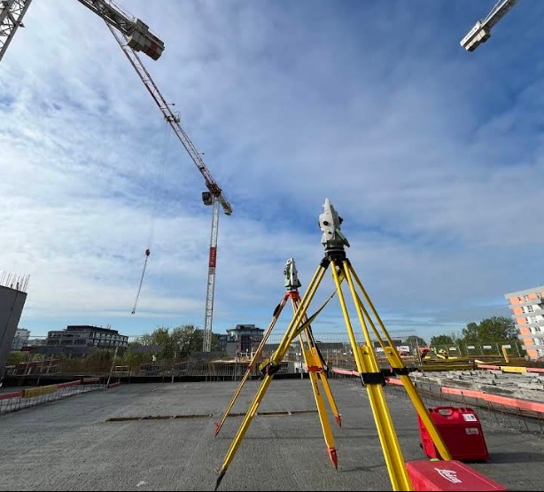 Dwa teodolity na budowie z żurawiami w tle, wykonujące pomiary geodezyjne na betonowej płycie. Czerwona skrzynka narzędziowa w prawym dolnym rogu.