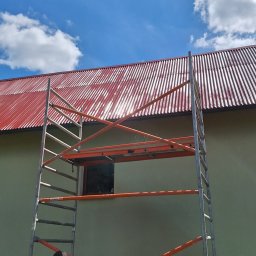 Paweł ZaborowskiDachy - Pracownik w rękawicach przygotowuje się do pracy na rusztowaniu przed budynkiem z czerwonym dachem z blachy trapezowej, widoczne niebieskie niebo z chmurami.