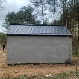 Paweł ZaborowskiDachy - Niewielki, szary budynek z nowym, czarnym dachem z dachówki bitumicznej w kształcie rybiej łuski, widoczny na tle lasu iglastego i pochmurnego nieba. W tle sylwetka człowieka w ciemnym ubraniu.