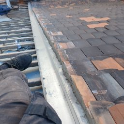 Paweł ZaborowskiDachy - Widok z góry na fragment dachu z częściowo ułożoną dachówką, widoczna metalowa obróbka blacharska i noga robotnika w czarnym obuwiu roboczym, w tle zabudowa miejska.